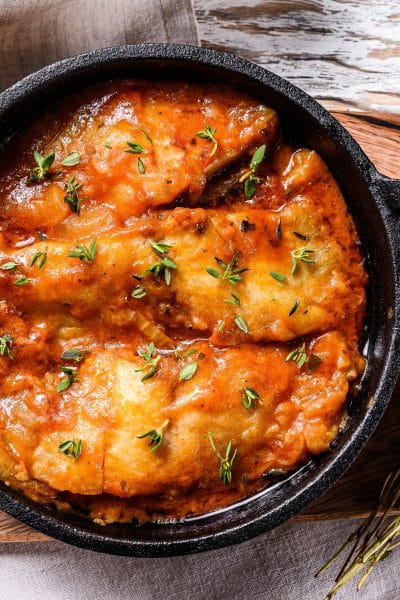 Baked halibut fish in a pan with tomato sauce. White wooden background. Top view